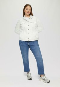 Witte denimjas met voorklessen, zilveren knopen; gecombineerd met blauwe rechte jeans en zilveren sportschoenen. Vlakke textuur, casual ontwerp.