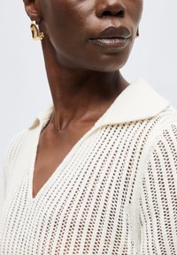 Άτομο με σκουρόχρωμο δέρμα που φοράει χρυσά αφηρημένα σκουλαρίκια και λευκό πουλόβερ ανοιχτής πλέξης με φαρδιά γιακά και V-λαιμόκοψη.