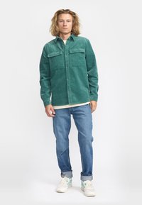 Groene corduroy button-up shirt met twee borstzakken, gedragen over een lichtgekleurde top en gecombineerd met blauwe spijkerbroek en witte sneakers.