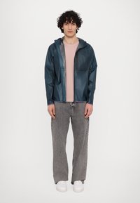 Gennemsigtig teal jakke med hætte, frontlynlås og brystlomme, parret med en lyserød t-shirt og grå brede jeans. Hvide sneakers.