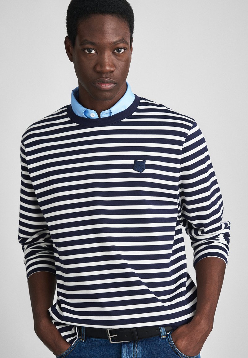 Navy and white striped long-sleeve shirt with a crew neck, featuring a small embroidered emblem on the chest and a blue collared shirt underneath.