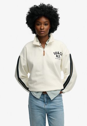 Sweatshirt crème à demi-zip avec des rayures bleu foncé sur les manches, logo brodé et texture douce. Associé à une chemise rayée en dessous.