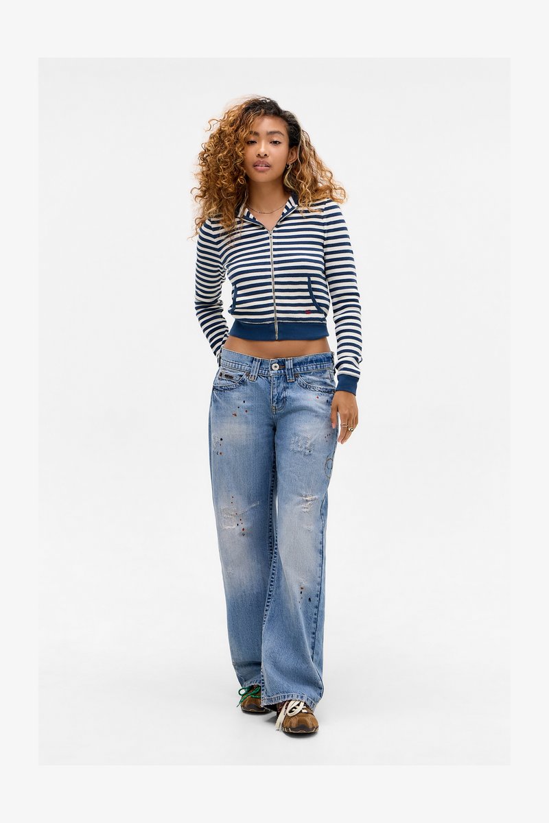 Jonge vrouw met krullend haar die een gestreepte zip-up cropped trui, versleten wijde jeans en bruine sneakers draagt, staande op een witte achtergrond.