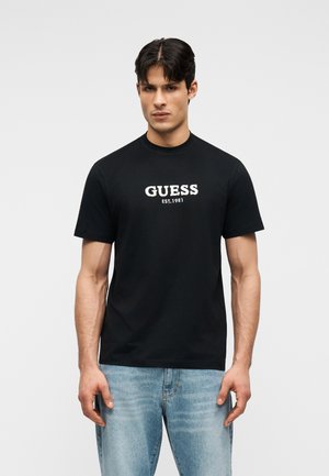 Jeune homme portant un T-shirt noir GUESS avec l'inscription "EST. 1981", associé à un jean bleu clair, se tenant devant un fond clair uni.