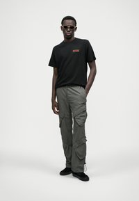 T-shirt noir avec lettrage orange, associé à un pantalon cargo vert olive comportant plusieurs poches. Des chaussures noires et des lunettes de soleil claires complètent le look.