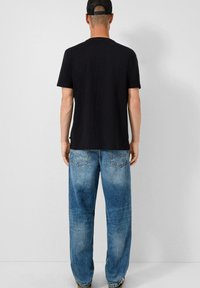 Juodas medvilninis marškinėliai su apvaliu kaklu, derinami su šviesiai mėlynomis, laisvai krentančiomis džinsinėmis kelnėmis, turinčiomis užpakalines kišenes ir išblukusiu tekstūru.
