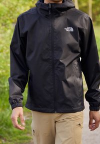 Veste imperméable noire avec fermeture éclair frontale, capuche ajustable, poignets élastiques, et un logo sur le côté gauche de la poitrine. Portée avec un pantalon beige.