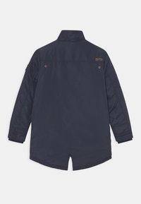 Námořnická modrá, dlouhá lehká bunda s vysokým límcem, prošívaným vzorem na rukávech a bočním rozparkem na zadní straně. Obsahuje nenápadné branding.