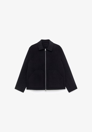 Schwarze Wolljacke mit geradem Schnitt, ausgestattet mit einem kragenlosen Ausschnitt, einem Frontreißverschluss und zwei großen Aufsetztaschen. Glatte Textur.