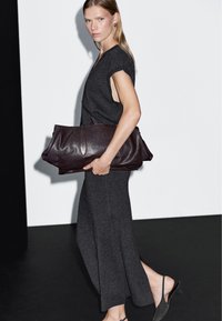 Vestito grigio scuro con maniche corte e spacchi laterali. Viene tenuta una grande borsa in pelle bordeaux scuro, caratterizzata da dettagli cuciti delicati.