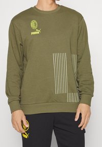 Sweat-shirt vert olive avec un design graphique composé de rayures blanches verticales. Comprend le logo Puma et un emblème de l'AC Milan sur la poitrine.
