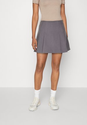 Kvinde iført et gråt plisseret skørt, beige top med korte ærmer, hvide sokker og lyse sneakers, der står mod en ensfarvet baggrund.