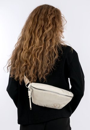 Osoba o długich, kręconych włosach nosi białą, fakturowaną torbę na ramię na czarnej kurtce, na tle jednolitej białej ściany.