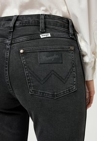 Jeans de mezclilla negra con una cintura alta, que presentan un parche de cuero y una costura distintiva en los bolsillos traseros, acompañados de herrajes plateados.