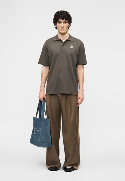 Jeune homme aux cheveux bouclés portant un polo marron et un pantalon large marron, tenant un sac fourre-tout bleu avec une écriture blanche.