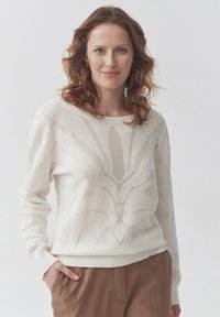 Suéter crema claro de manga larga, con un patrón floral texturizado. Puños y dobladillo acanalados, combinado con pantalones marrones.