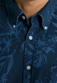 Detailní záběr na osobu v námořnické modré košili na knoflíky s květinovým vzorem a bílými knoflíky, která ukazuje část krku a límce.