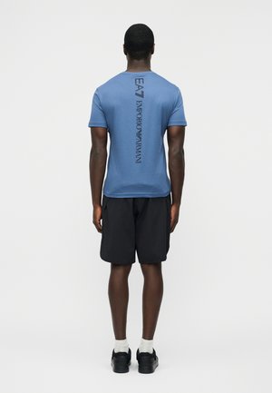 Mężczyzna ubrany w niebieską koszulkę Emporio Armani z pionowym czarnym napisem logo na plecach, czarne szorty, białe skarpetki i czarne buty, stojący.