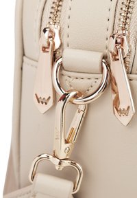 Beige kabelka zblízka s dvojitými zipy, růžovým zlatým kováním, oválným kovovým kroužkem a koženými detaily. Hladký povrch.