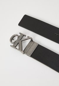 Črn usnjen pas z zaponko iz srebrne tone, zasnovano v obliki stiliziranega logotipa "CK". Gladka tekstura, minimalen dizajn.