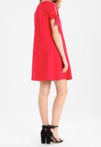 Robe rose à manches courtes avec des découpes aux épaules, silouhette évasée et texture lisse. Associée à des chaussures à talons hauts noires.