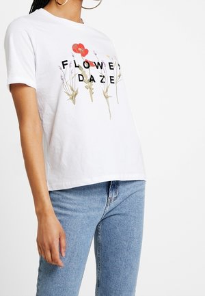 Vrouw draagt wit T-shirt met rode klaproos en tekst "FLOWER DAZE", gecombineerd met lichtblauwe spijkerbroek tegen een witte achtergrond.