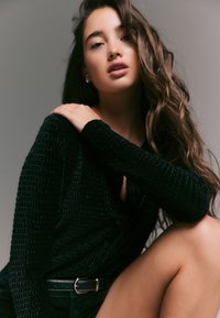 Camicia nera con bottoni e trama strutturata, maniche lunghe, indossata sopra pantaloncini scuri. La modella ha capelli mossi e sciolti e una cintura argentata che evidenzia la vita.