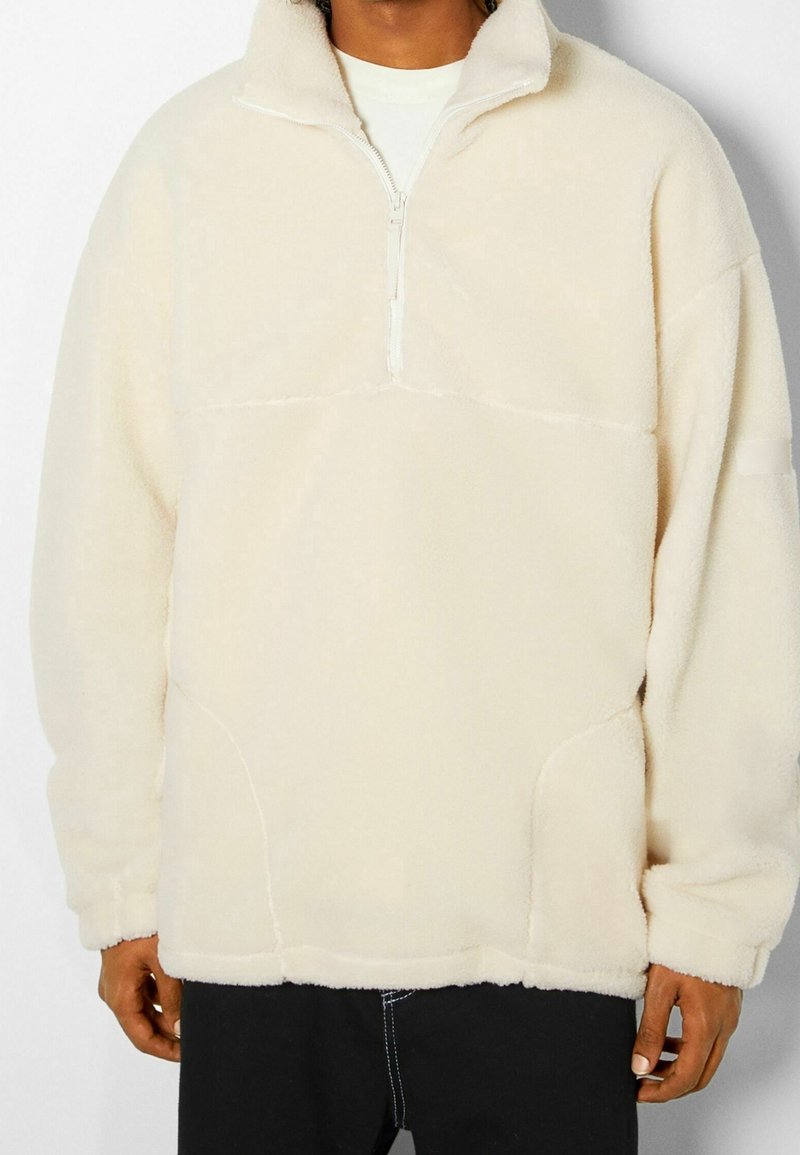 Fleece-pullari vaaleanbeige värissä, jossa on neljänneksen vetoketju edessä, korkea kaulus ja sivutaskut. Pehmeä rakenne, jossa on ommeltuja yksityiskohtia.