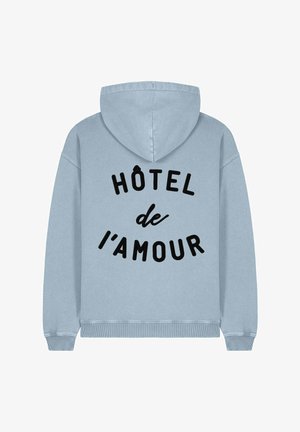 Hoodie u svijetloplavoj boji, s crnim tiskanim natpisom "HÔTEL de l'amour" na stražnjoj strani. Mekani materijal s rebrastim rubovima.