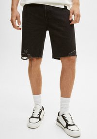 Pantalones cortos de mezclilla negros con dobladillos deshilachados y detalles desgastados, combinados con calcetines blancos y zapatillas en blanco y negro. Diseño minimalista.