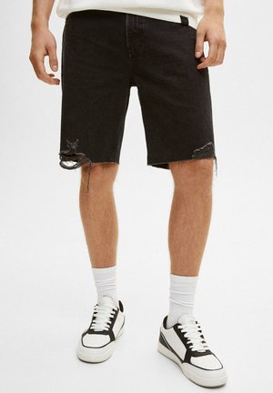 Svarta jeansshorts med fransade kanter och slitna detaljer, kombinerade med vita strumpor och svart-vita sneakers. Minimalistisk design.