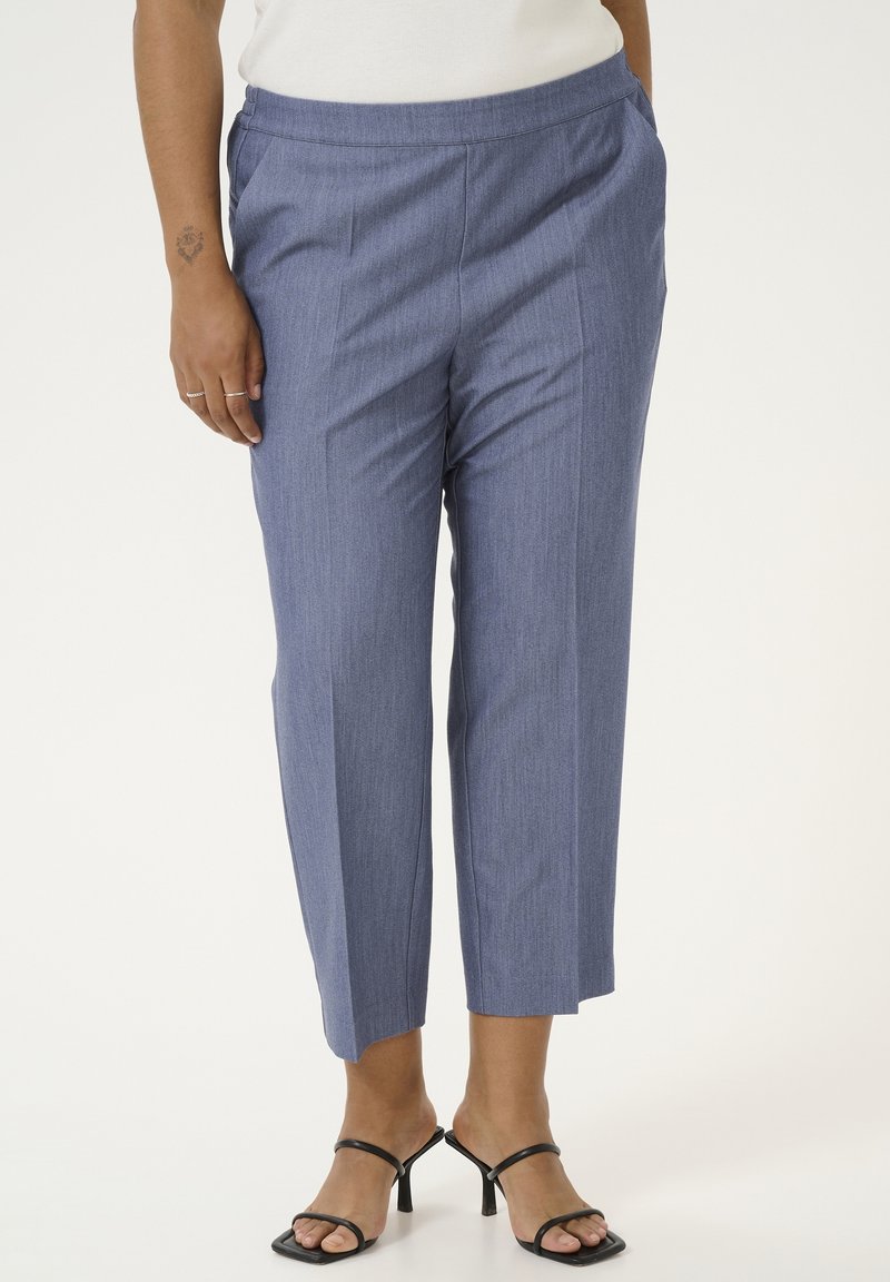 Femme portant un pantalon bleu taille haute court avec des plis et des sandales noires à talons, debout devant un fond clair uni.