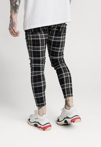 Jeans skinny a quadri neri con accenti rossi e gialli, realizzati in materiale elastico. Abbinati a sneakers chunky bianche con dettagli neri e rossi.