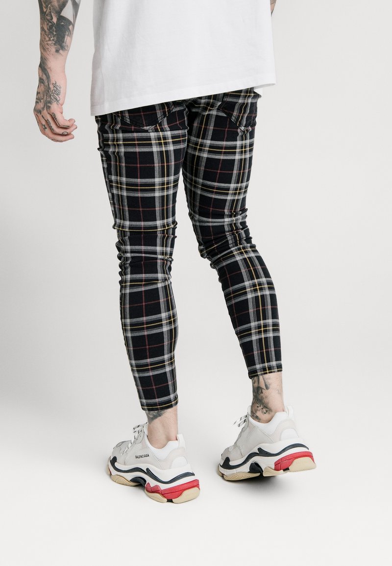 Jeans skinny a quadri neri con accenti rossi e gialli, realizzati in materiale elastico. Abbinati a sneakers chunky bianche con dettagli neri e rossi.