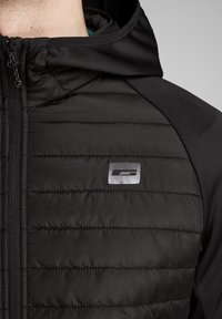 Nahaufnahme einer schwarzen gesteppte Jacke mit Kapuze, die den Reißverschluss und ein kleines reflektierendes rechteckiges Logo auf der Brust zeigt.