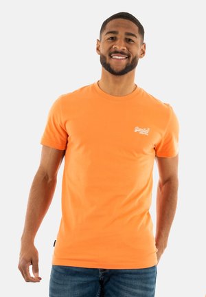 Hombre sonriente con camiseta naranja de manga corta con texto pequeño blanco en el pecho y jeans azules, de pie frente a un fondo liso.