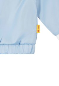 Veste bleu clair en tissu lisse, dotée de poignets élastiques et d'une étiquette de marque jaune sur la manche. Finie en blanc.