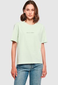 Mintgrøn bomulds t-shirt med en afslappet pasform, korte ærmer og et lille logo, der står "Flutter & Bloom" centreret på brystet.