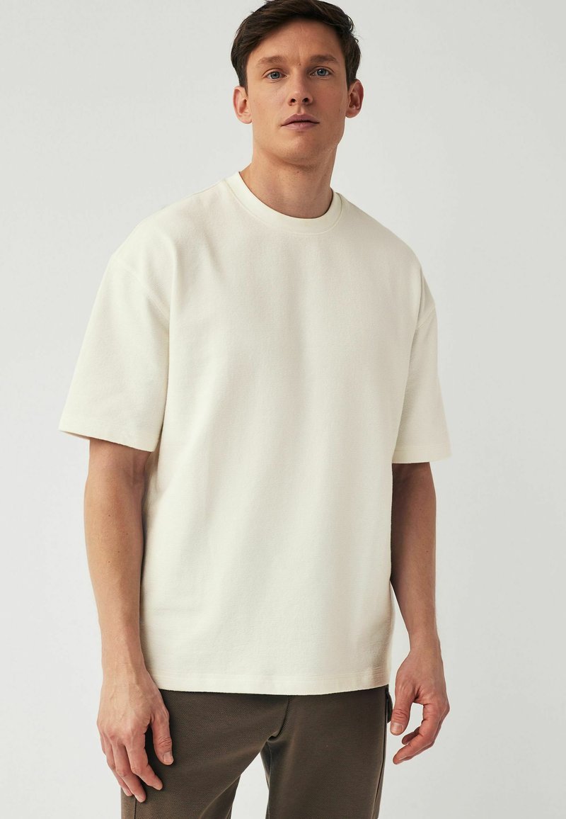 T-shirt crema a maniche corte realizzata in un tessuto testurizzato, caratterizzata da un colletto rotondo e una vestibilità ampia. Abbinata a pantaloni scuri.