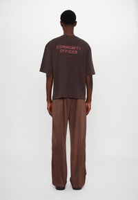 Brun oversized t-skjorte med teksten "SAMFUNNSFUNKSJONÆR" på baksiden, sammen med brune vide bukser. Glatt stoff, avslappet passform.