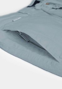 Tissu bleu clair avec une poche zippée partiellement ouverte, montrant une tirette de fermeture éclair visible et la texture du tissu.