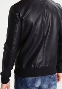 Giacca bomber nera in pelle con texture liscia, polsini e orlo a coste, caratterizzata da un design pulito sul retro e nessun hardware visibile.
