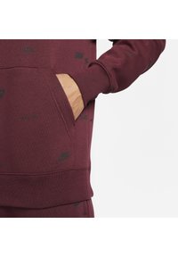 Burgundy sweatshirt with a front pocket, featuring black Nike logos in a repeated pattern. Made of soft fabric with ribbed cuffs.