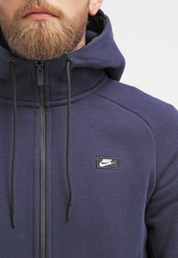 Tmavě modrá mikina na zip s vysokým límcem, �šňůrkami na stažení a černou nášivkou s logem Nike na prsou. Vyrobena z texturované látky.