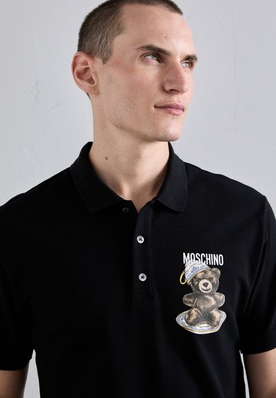 Svart polo-tröja med en texturerad yta, prydd med en björngrafik som bär en hatt, och texten "Moschino", kompletterad med silverknappar.