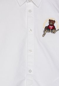 Witte katoenen blouse met een berenborduursel, de tekst "Opgericht in 1981" en een logo in rood en zwart. Knopen zijn van doorzichtig plastic.