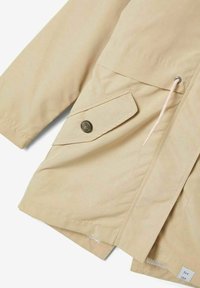Manga y parte delantera de una chaqueta beige que muestra un bolsillo con solapa abotonado y un cordón blanco con ojales de metal en una tela ligera.