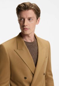 Blazer cammello doppiopetto con rever a punta, realizzato in un tessuto morbido, abbinato a un maglione marrone a trama sottostante.