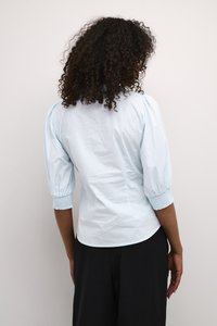 Blusa azzurra chiara con maniche a sbuffo, caratterizzata da un design attillato e polsini elasticizzati. Texture liscia e stile minimalista.