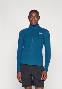 Chaqueta de forro polar en color verde azulado con un alto cuello, cremallera completa y dos bolsillos laterales. Logotipo destacado en la zona del pecho izquierdo. Combinada con pantalones cortos negros.